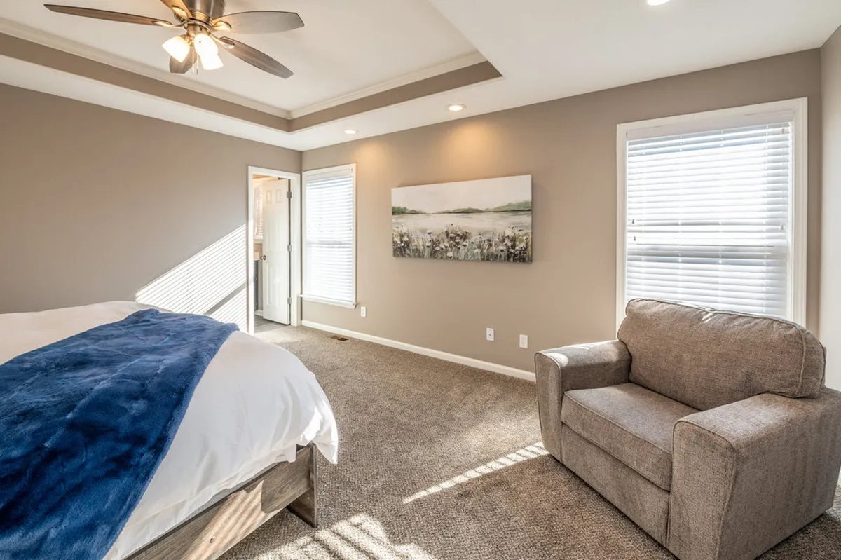A tranquil bedroom scene with a comfortable bed draped in a blue throw blanket, an inviting armchair, and a landscape painting above. The space is bathed in natural light from a window, fostering a peaceful and cozy atmosphere.