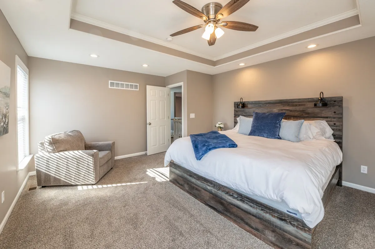 A serene bedroom featuring a plush bed adorned with soft bedding and an armchair nestled near a large window. Natural light filters gently into the space, illuminating calming décor and a ceiling fan above, inviting relaxation and rest.