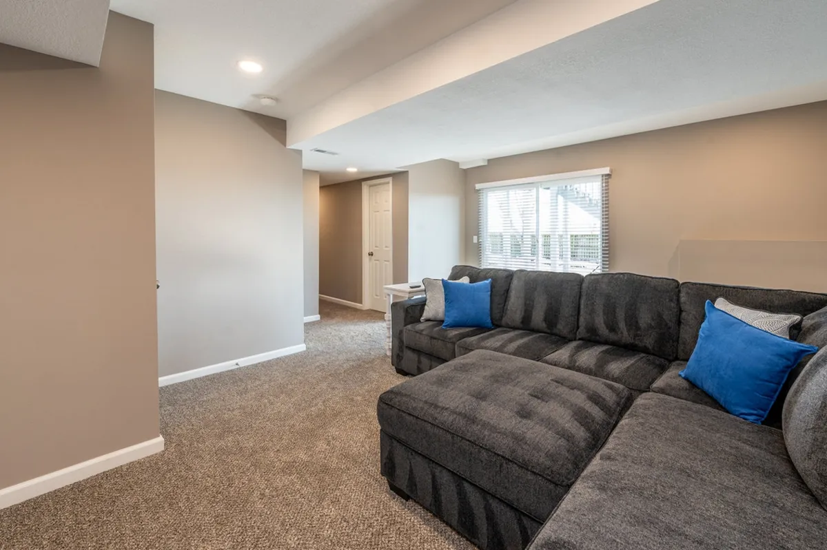 A light-filled living area with a sectional sofa perfectly positioned near expansive windows. The windows flood the room with natural light, highlighting the plush carpet and offering tranquil views, making it an ideal spot for reading or relaxing.