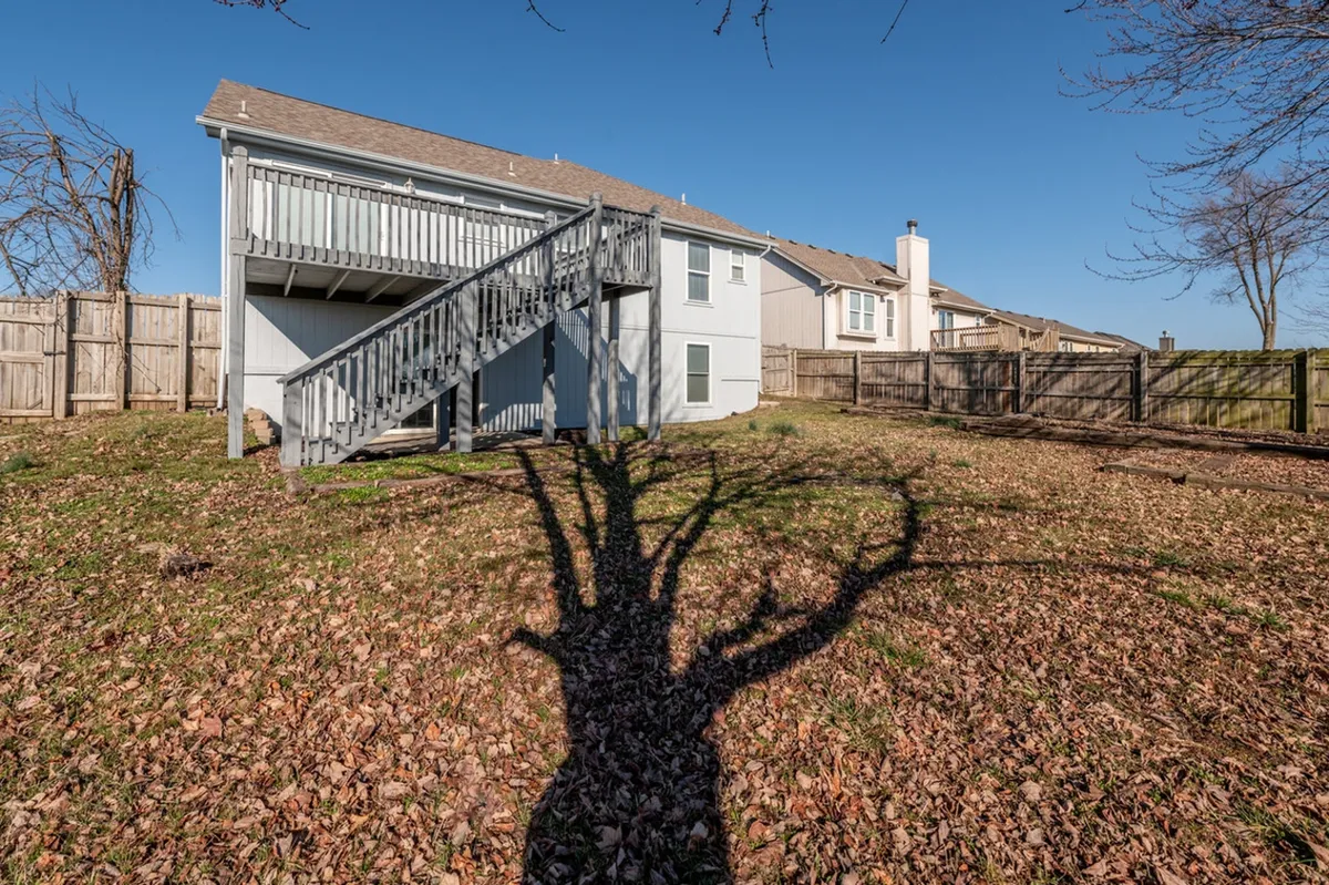 This exterior image displays the home’s façade, a staircase leading to the entry, and the soft shadow of a tree across the yard. The sun-dappled setting suggests a tranquil outdoor environment, with hints of green space ideal for strolling or relaxing near the home.