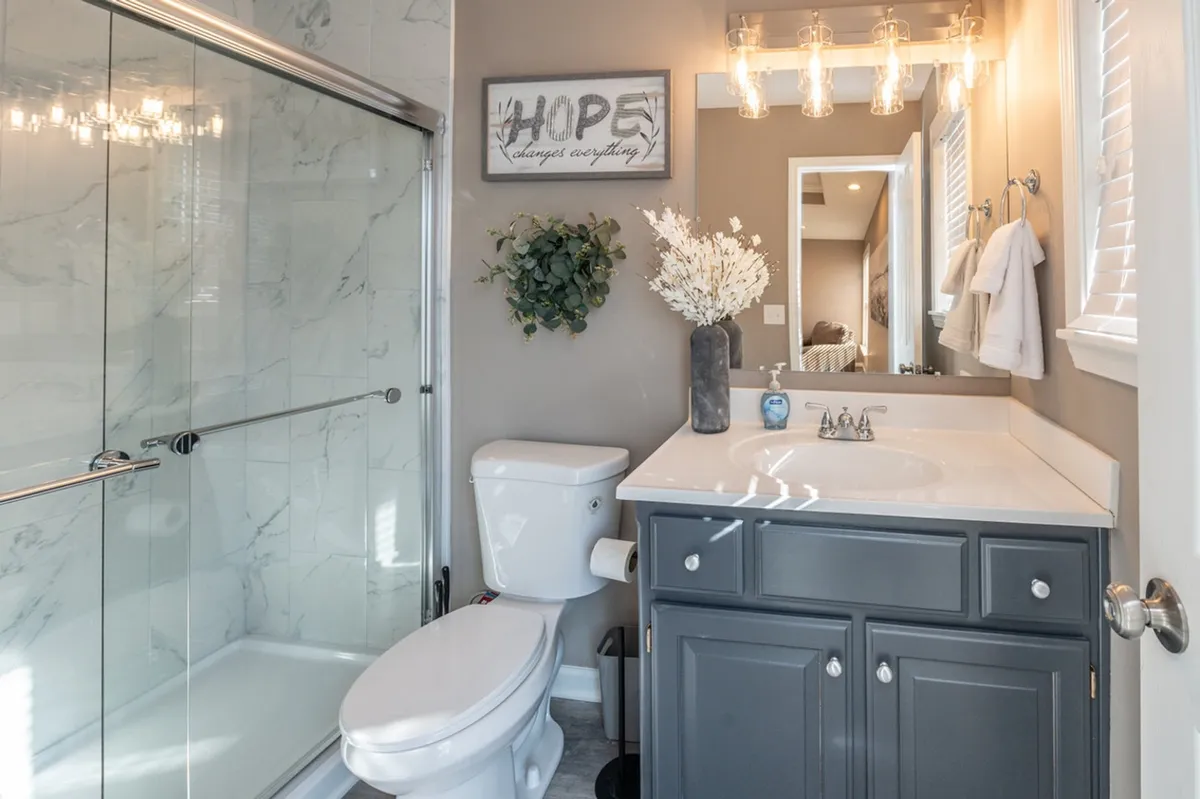A modern bathroom featuring a pristine shower with glass doors, a sleek vanity with dark cabinetry, and a decorative vase with flowers next to a framed 'HOPE' sign. The space feels uplifting, organized, and conducive to self-care routines.