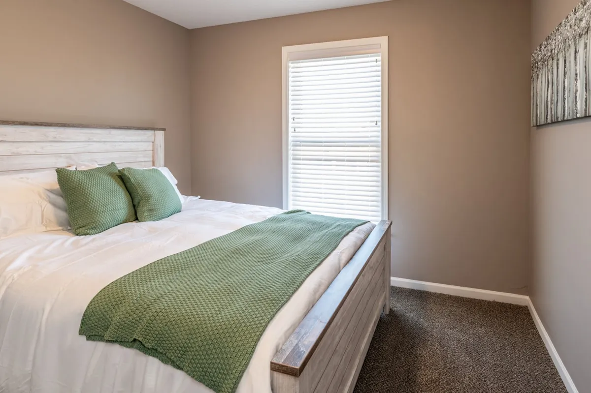 A sunlit bedroom with a plush bed covered in a green throw blanket faces a window, inviting natural light to stream in. The simple, uncluttered décor emphasizes peacefulness, and outside the window, hints of nature suggest a connection to the lakeside environment.